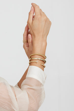 A hand wears the Kinsey Designs Mara Cuff—a gold plated adjustable bracelet—styled with gold bangles, a ring, and a sheer white sleeve, set against a plain background.