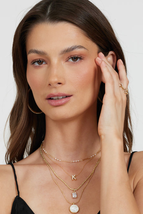 Woman with wavy brown hair wearing layered Kinsey Designs gold necklaces, including a Marina Pearl Necklace, hoop earrings, and a black top, touching her face.