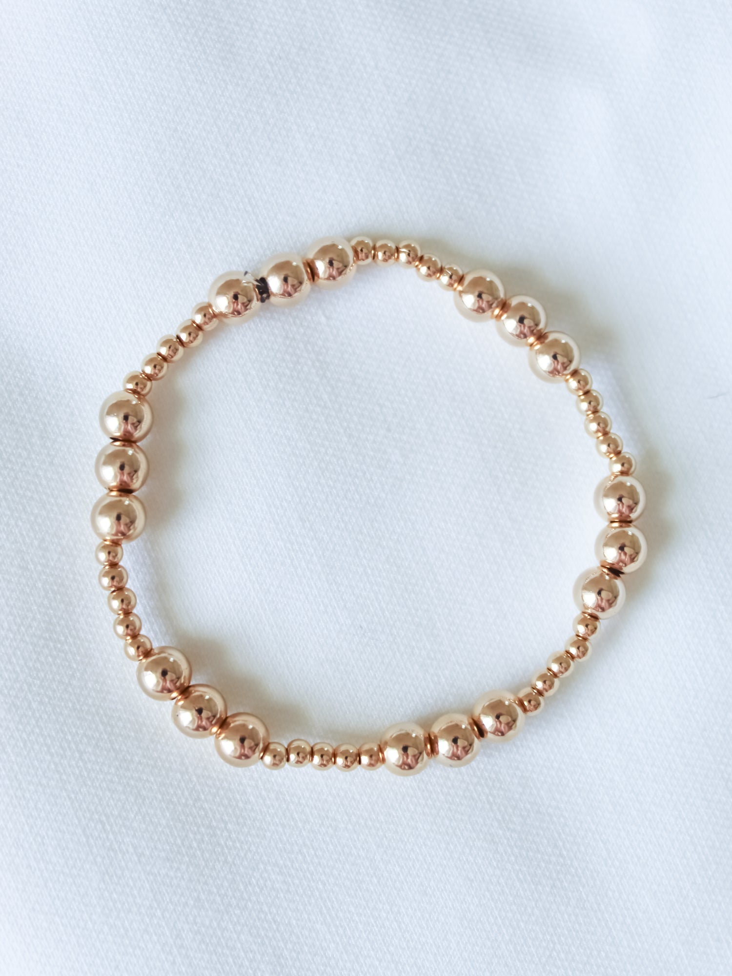 The Mia Bracelet by Kinsey Designs, shown in multiple sizes, features gold-filled beads and an adjustable stretch design, arranged in a circle on a white fabric background.