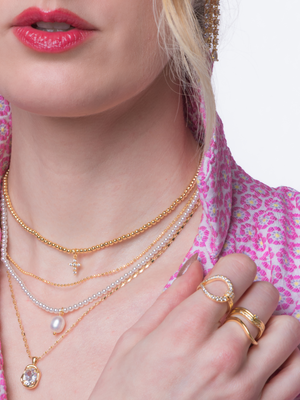 A woman wears layered gold and pearl necklaces, Kinsey Designs' Miller Curve Ring, and stackable rings on her hand, styled with a pink patterned blouse—close-up of her neck and hand.