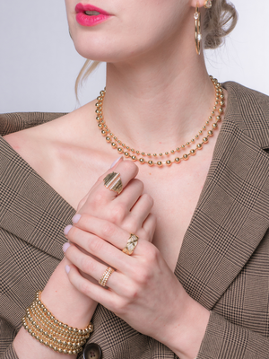 A woman in a plaid blazer wears gold jewelry—including the adjustable Kinsey Designs Nenya Ring—paired with a necklace, bracelet, hoop earrings, a leaf ring, and pink lipstick.
