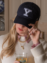 A woman with long blonde hair wears a Yale cap, cream sweater, and gold Kinsey Designs Numerical Slide charm necklace, paired with gold jewelry, as she holds a glass indoors.