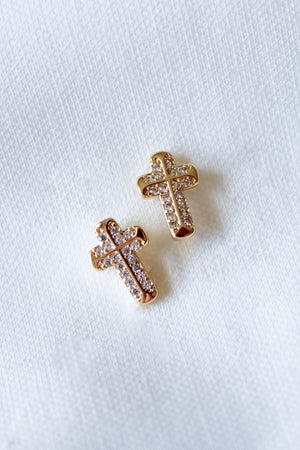 The Noah Cross Studs by Kinsey Designs feature two gold cross earrings adorned with small clear gemstones on a white fabric background, ideal for those who love minimalist or meaningful faith jewelry.