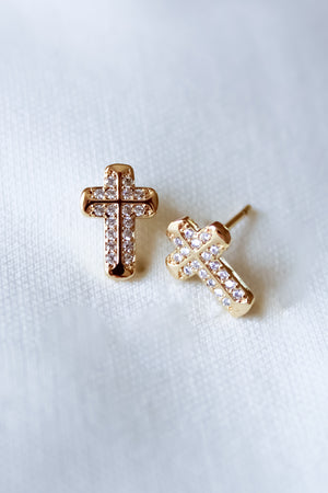 The Noah Cross Studs by Kinsey Designs feature gold cross earrings with small clear stones on a soft white fabric background. These minimalist earrings add timeless, subtle elegance and a touch of faith to any look.