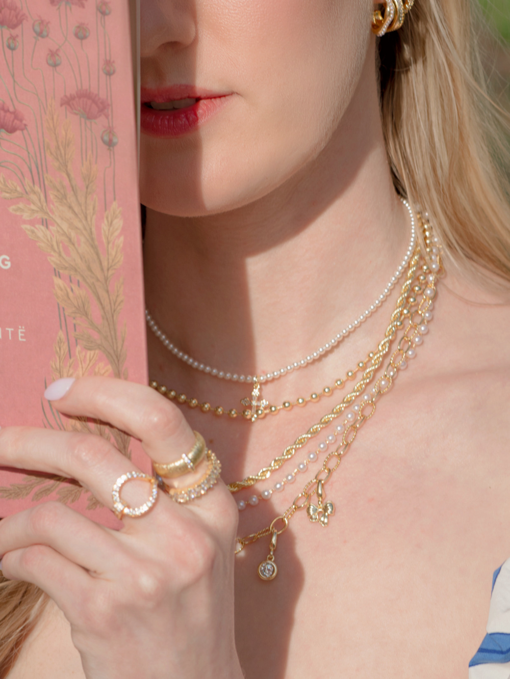 A woman holds a pink floral book, wearing the Olivia Layer Necklace by Kinsey Designs—layered chains with pearls and gold—partially covering her face.