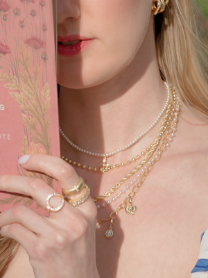 A woman holds a pink floral book, wearing the Olivia Layer Necklace by Kinsey Designs—layered chains with pearls and gold—partially covering her face.