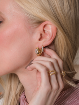 A blonde woman wearing gold hoop earrings, featuring the Kinsey Designs Pave Huggie Earring, along with gold rings, touches her ear.