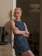 A woman in a blue plaid dress stands with arms crossed, leaning against a kitchen counter and looking to the side. Her Perry Braided Knot Earrings by Kinsey Designs add a modern touch to her look.