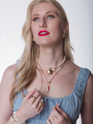 A woman with long blonde hair and red lipstick, wearing a blue dress and the Kinsey Designs Paulette Puffy Star Necklace—a layered gold filled piece with a star pendant—looks upward.