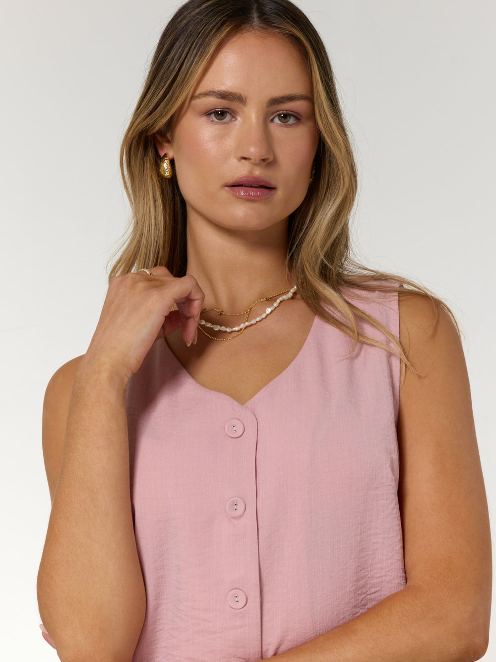 A woman with wavy blonde hair wears a pink sleeveless button-up top and the Kinsey Designs Pearl Layering Necklace, posing against a white background.