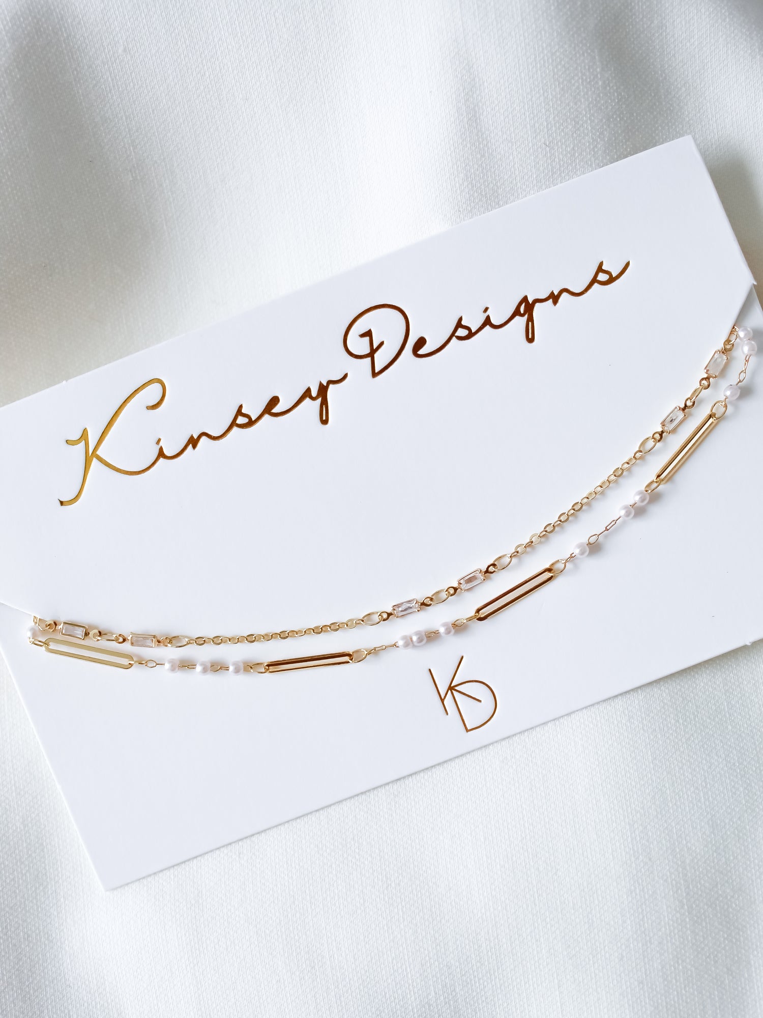 The gold and pearl chain bracelet is displayed on a Kinsey Designs card atop a white fabric background—perfect for pairing with the Kinsey Designs Pelly Layer Necklace from our exclusive sample sale.