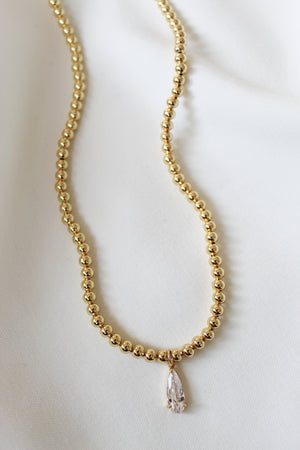 The Penn Necklace by Kinsey Designs Archives, featuring gold beads and a pear-shaped pendant, is displayed elegantly on a white fabric background.
