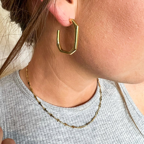 A woman wears the Kinsey Designs Archives Polly Hoop, a gold plated geometric earring (1" wide, 1.25" long), paired with a delicate gold chain necklace and a gray ribbed top.