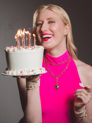 A woman wearing the Kinsey Designs Poppy Puffy Heart Necklace holds a cake with candles.