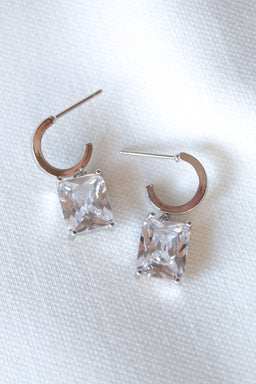 Kinsey Designs’ Prism Huggies showcase square princess-cut CZ gemstones, elegantly displayed on a white textured fabric background.