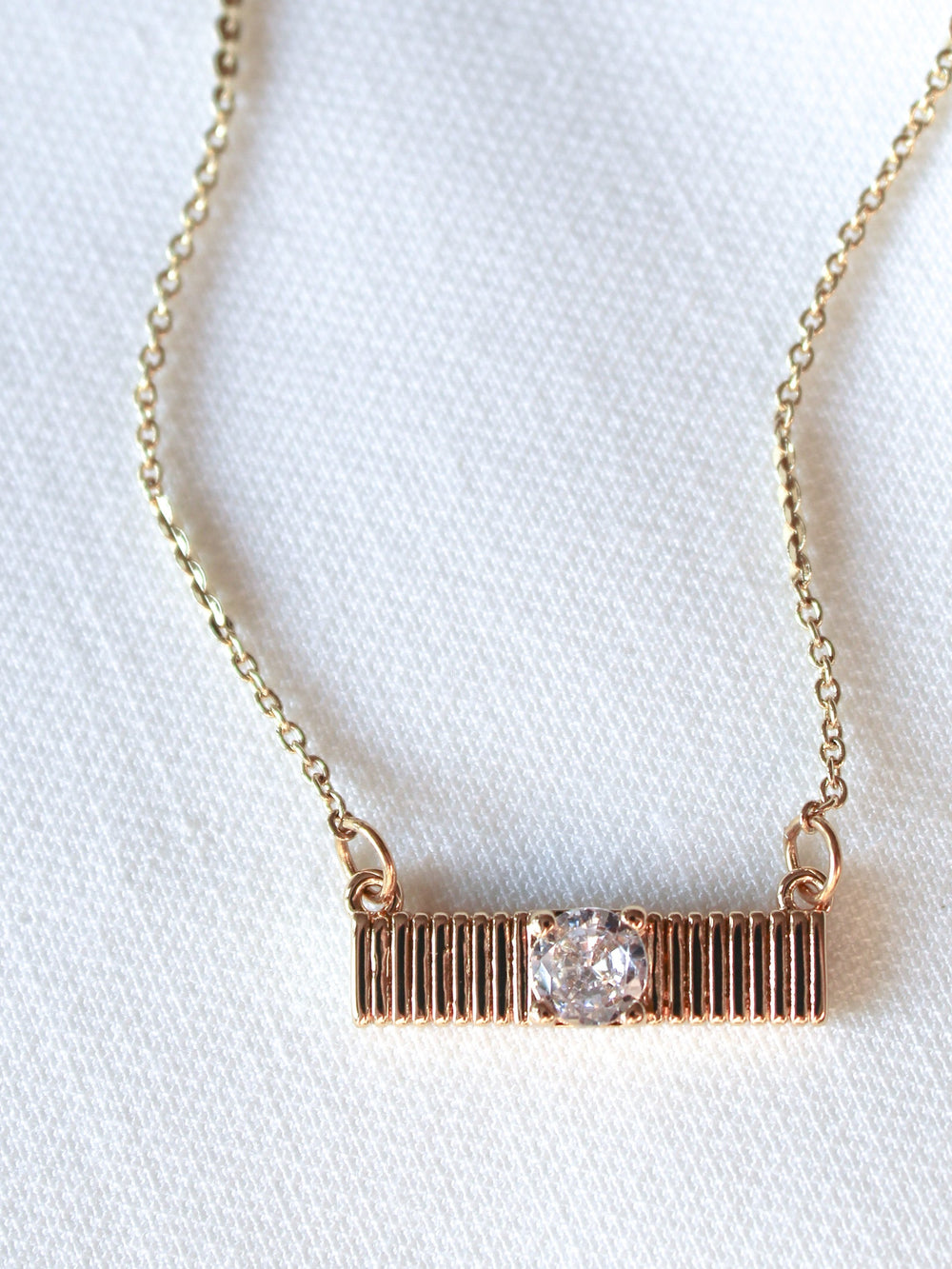 The Quinn Bar Necklace by Kinsey Designs showcases a horizontal gold bar pendant with a central clear gemstone, elegantly displayed on light fabric—perfect as a jewelry gift for any occasion.