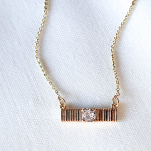 The Quinn Bar Necklace by Kinsey Designs showcases a horizontal gold bar pendant with a central clear gemstone, elegantly displayed on light fabric—perfect as a jewelry gift for any occasion.