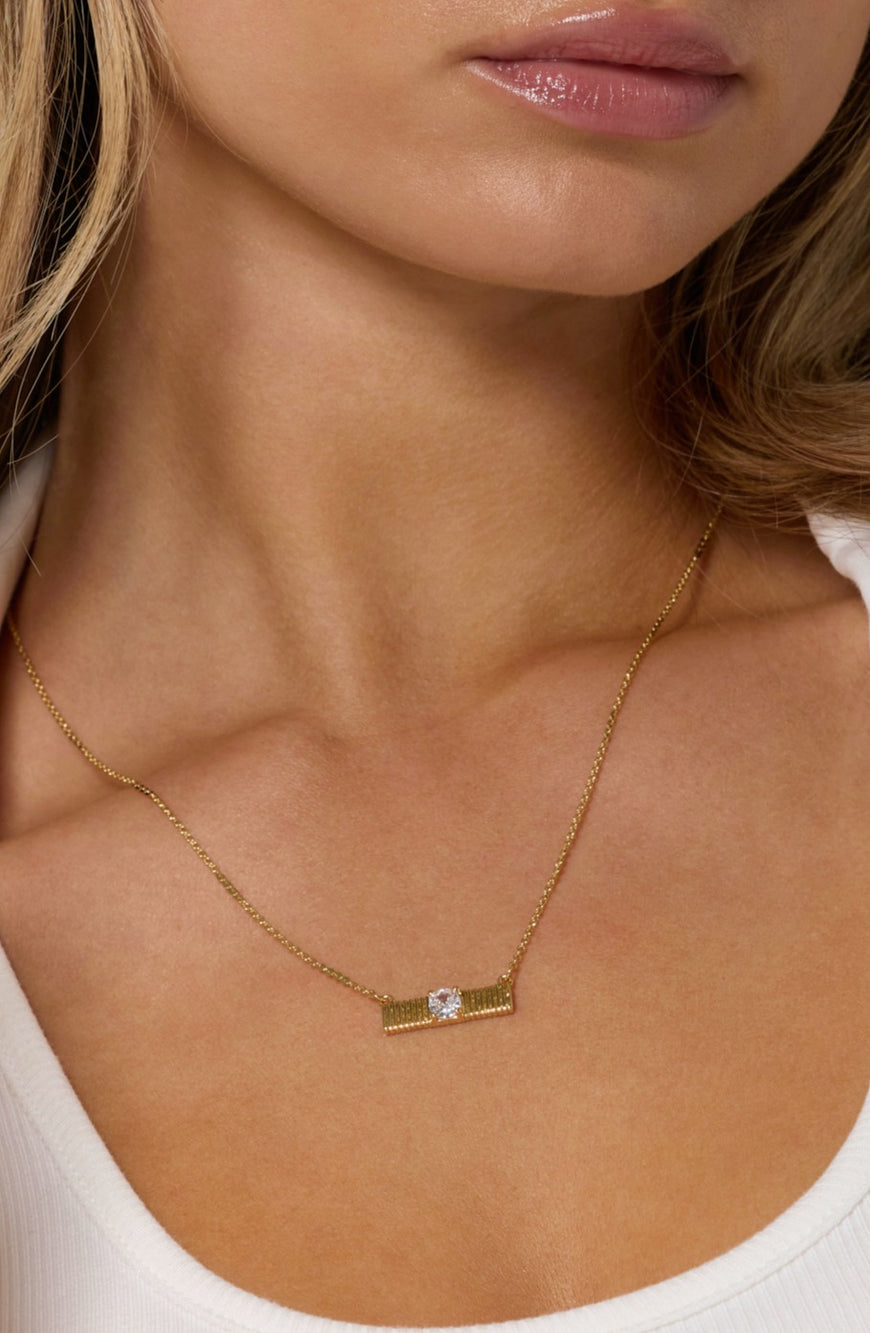 A woman wears the Kinsey Designs Quinn Bar Necklace, a gold pendant with a rectangular design and central clear gemstone—an elegant gift for any occasion.