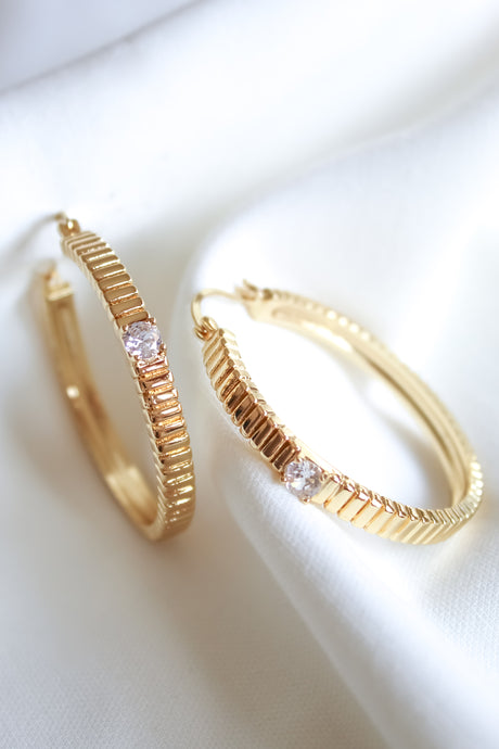 The Quinn Hoop Large by Kinsey Designs Archives features a textured gold filled design with a small clear gemstone, displayed on white fabric—perfect for adding elegance to any look. Shop exclusive sample sale finds now.