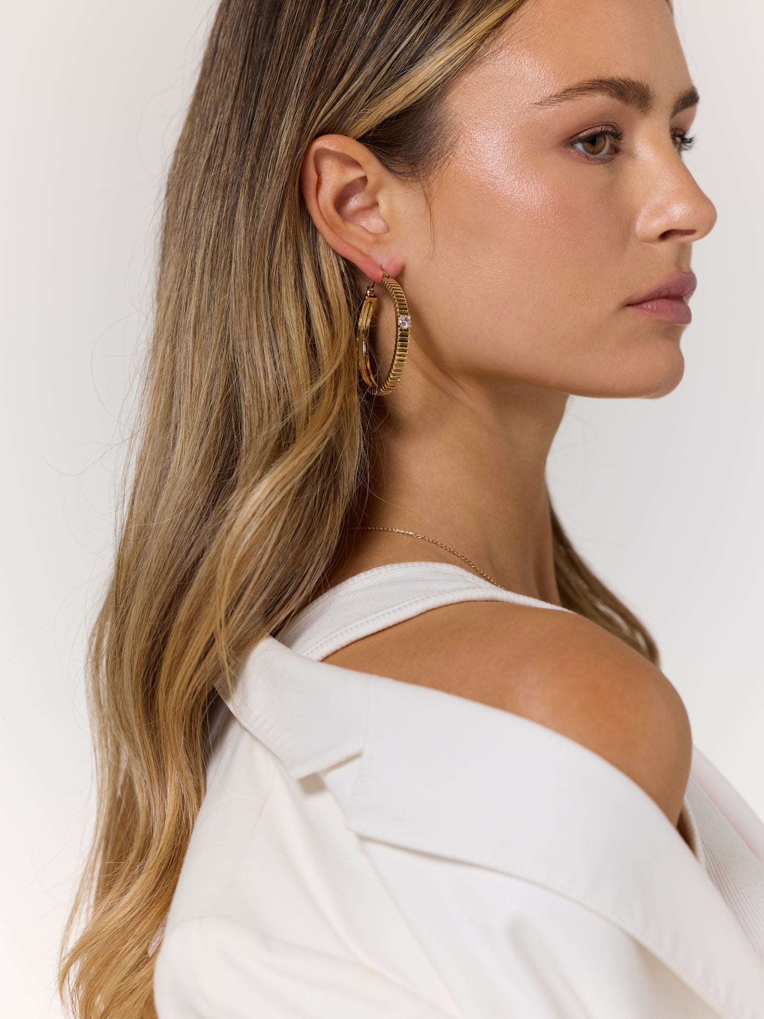 Woman with long blonde hair, wearing a white top and a white jacket off her shoulder, accessorizes with Kinsey Designs Archives’ Quinn Hoop Large gold-filled earrings.