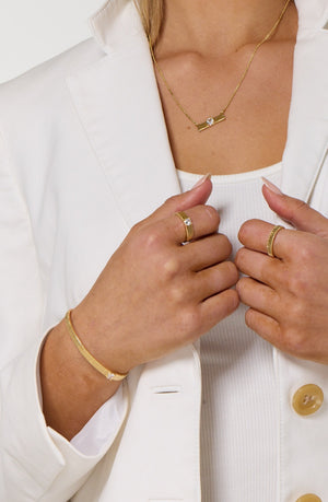 A woman in a white blazer showcases gold jewelry near her chest, featuring the Kinsey Designs Quinn Ring, a stacking band, CZ stone gold ring, necklace, and bracelet against a white background.