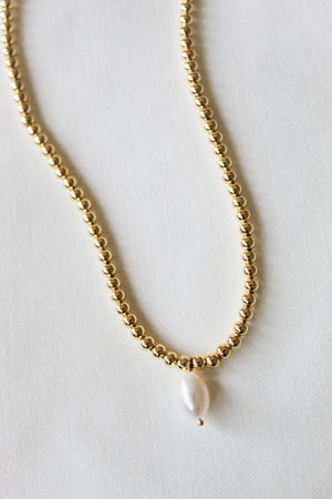 The Kinsey Designs Reed Necklace features a single white pearl pendant on a gold-filled chain, elegantly showcased against a white fabric backdrop.