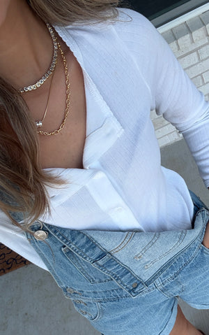 A woman in a white shirt and denim overalls stands on a light-colored surface, accessorized with the Josephine Link Necklace by Kinsey Designs.