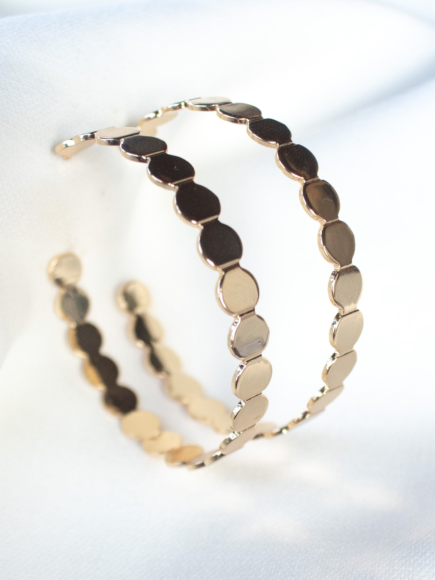 The Kinsey Designs Archives "Round and Round Hoop" earrings feature a scalloped, disc-like gold-filled design, measuring 2" wide and elegantly showcased on a soft white fabric background.
