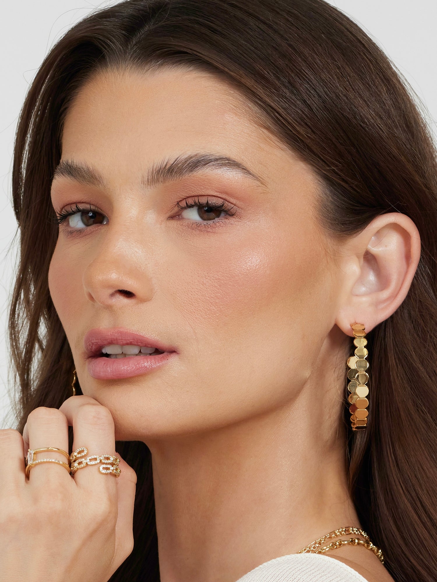 A woman with long brown hair gazes softly at the camera, wearing Kinsey Designs Archives' Round and Round Hoop gold-filled 2" earrings, paired with gold rings and a gold necklace.