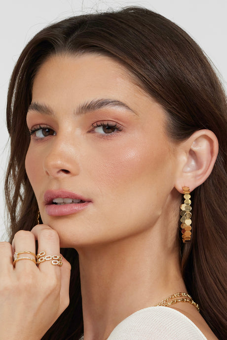 A woman with long brown hair gazes softly at the camera, wearing Kinsey Designs Archives' Round and Round Hoop gold-filled 2" earrings, paired with gold rings and a gold necklace.