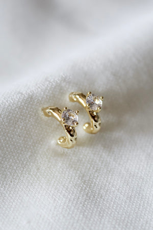 The Kinsey Designs Rugo Hoop earrings, crafted in gold and adorned with clear gemstones, are elegantly displayed on a soft white fabric background.