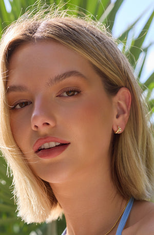 A blonde woman smiles outdoors in front of green plants, wearing Kinsey Designs’ Rugo Hoop gold-filled earrings.