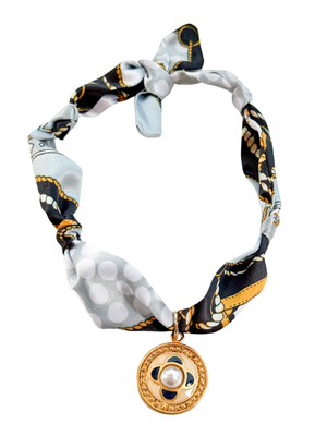 The Sassari Scarf Necklace by Kinsey Designs features a black, white, and gold pattern, tied in a knot and accented with a vintage pendant for an elegant look.