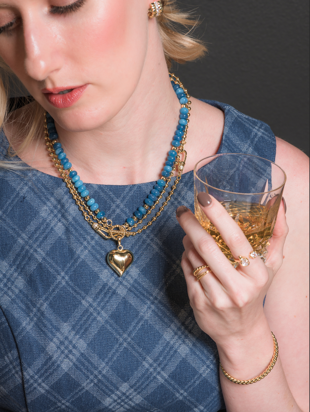 A woman in a blue plaid dress holds a drink, wearing gold jewelry and the Kinsey Designs Juniper Ball Chain Necklace with a heart pendant.