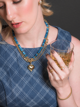 Woman in a blue plaid dress holds a glass of whiskey, accessorized with gold jewelry including the sparkling Kinsey Designs Lorelai Wrap Ring and a blue beaded necklace.