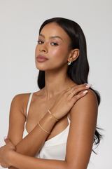 A woman with long dark hair wears the Kinsey Designs Nora Bracelet, gold plated, styled with a white top and arms crossed against a plain background.