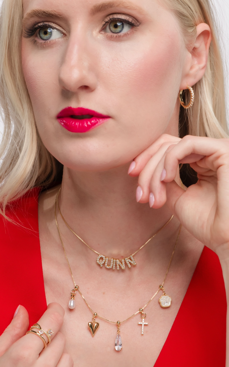 A woman wearing red lipstick and a gold "QUINN" name necklace, along with a charm necklace, is dressed in red. Her ensemble is completed with a Gold Filled Charm Bar Chains and a delicate Kinsey Charm Bar Vintage Heart Drop Charm.