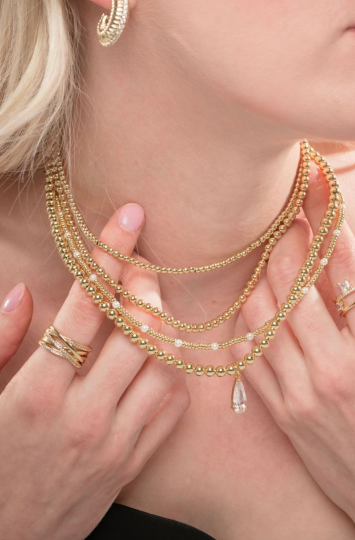 A woman wears the Kinsey Designs Archives Penn Necklace layered with gold beaded chains, paired with gold rings and hoop earrings.