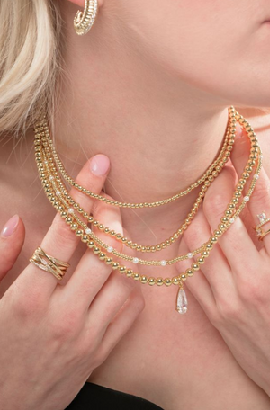 A woman wears the Kinsey Designs Archives Penn Necklace layered with gold beaded chains, paired with gold rings and hoop earrings.
