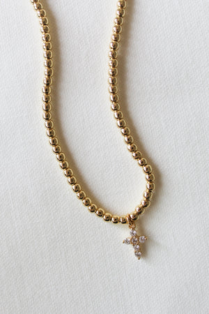 The Selena Necklace by Kinsey Designs features 3mm gold beads and a crystal-studded cross pendant, secured with a clasp and extender, elegantly displayed on a light textured surface.