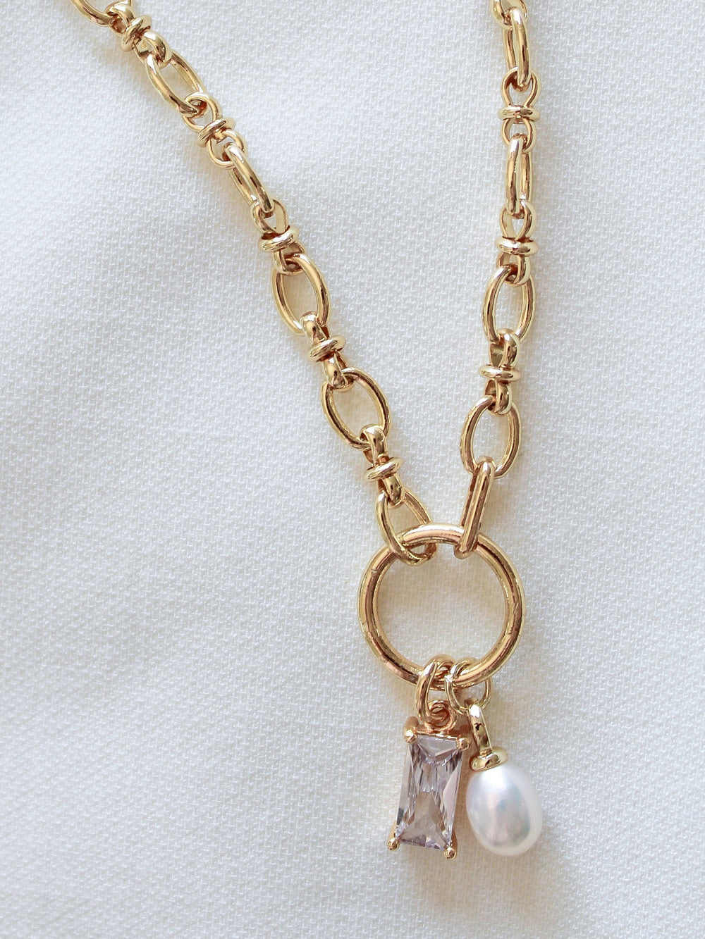 The Sophie Pearl Necklace by Kinsey Designs Archives features a gold-filled chain with a circular pendant, pearl charm, and rectangular cubic zirconia gemstone, elegantly displayed on white fabric.