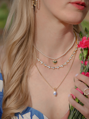 A woman shown from the shoulders up wears a pearl necklace layered with Kinsey Designs' Tilly Puff Heart Necklace and holds pink flowers.