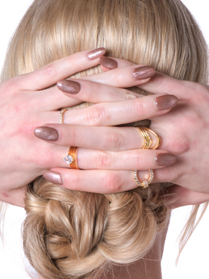 A woman with manicured nails and gold rings, including the Kinsey Designs Tally Triple Ring with CZ crystals, covers the back of her head with her hands; her hair is styled in a low bun.