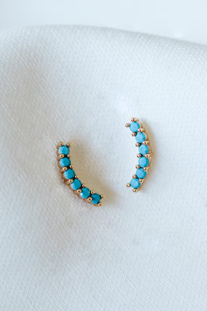 The Tide Climber earrings by Kinsey Designs, featuring small turquoise stones set in gold fill, are showcased on a soft white textured fabric—a stunning jewelry piece perfect for any sample sale.