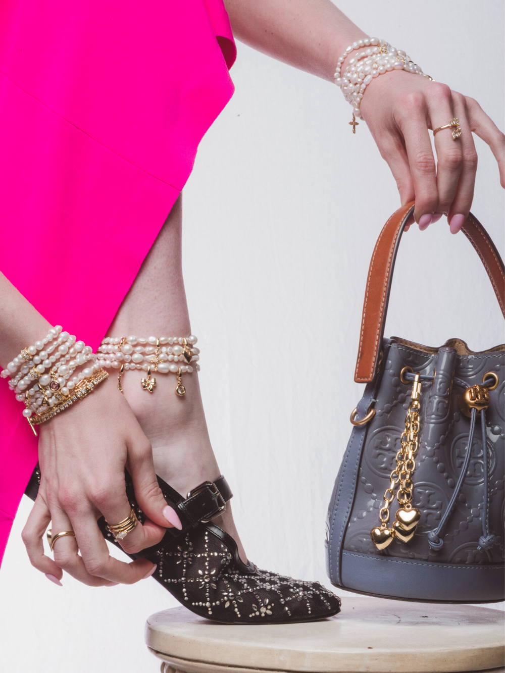A woman in a pink dress wears pearls, jeweled shoes, and holds a gray handbag featuring the Kinsey Designs Triple Heart Bag Charm on the table.