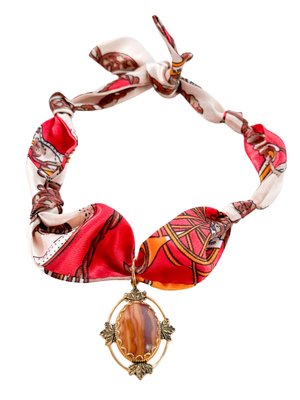 The Venice Scarf Necklace by Kinsey Designs features a red-patterned silk scarf and a vintage-inspired pendant with a gold oval gemstone and delicate leaf accents.