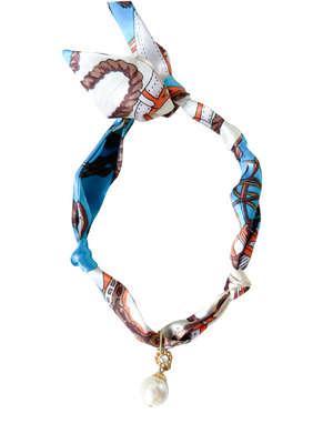 The Kinsey Designs Volterra Scarf Necklace features a colorful patterned scarf bracelet with a knot, a dangling pearl charm, and a gold fish pendant, displayed on a white background.
