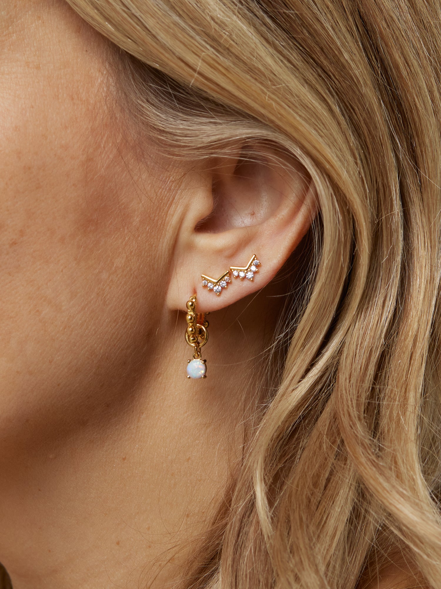 Close-up of a woman's ear with three gold filled earrings from Kinsey Designs Archives, featuring the Villa Stud and a dangling white stone. Add elegance—shop the Sample Sale now!.