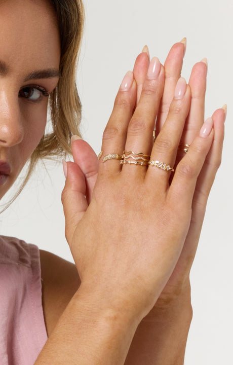 A woman with manicured nails wears multiple Kinsey Designs Venetia Rings, some adjustable, on her fingers near her face, adding versatility and elegance to her look.
