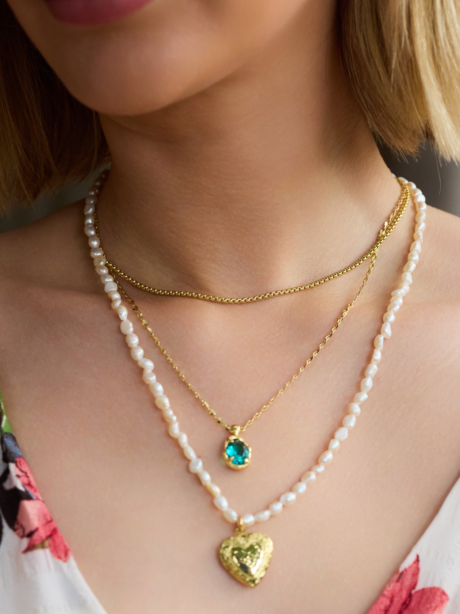 A woman wears three layered necklaces: an 18-inch gold filled chain, the Kinsey Designs Archives Birdie Blue Necklace, and a pearl necklace with a gold heart.